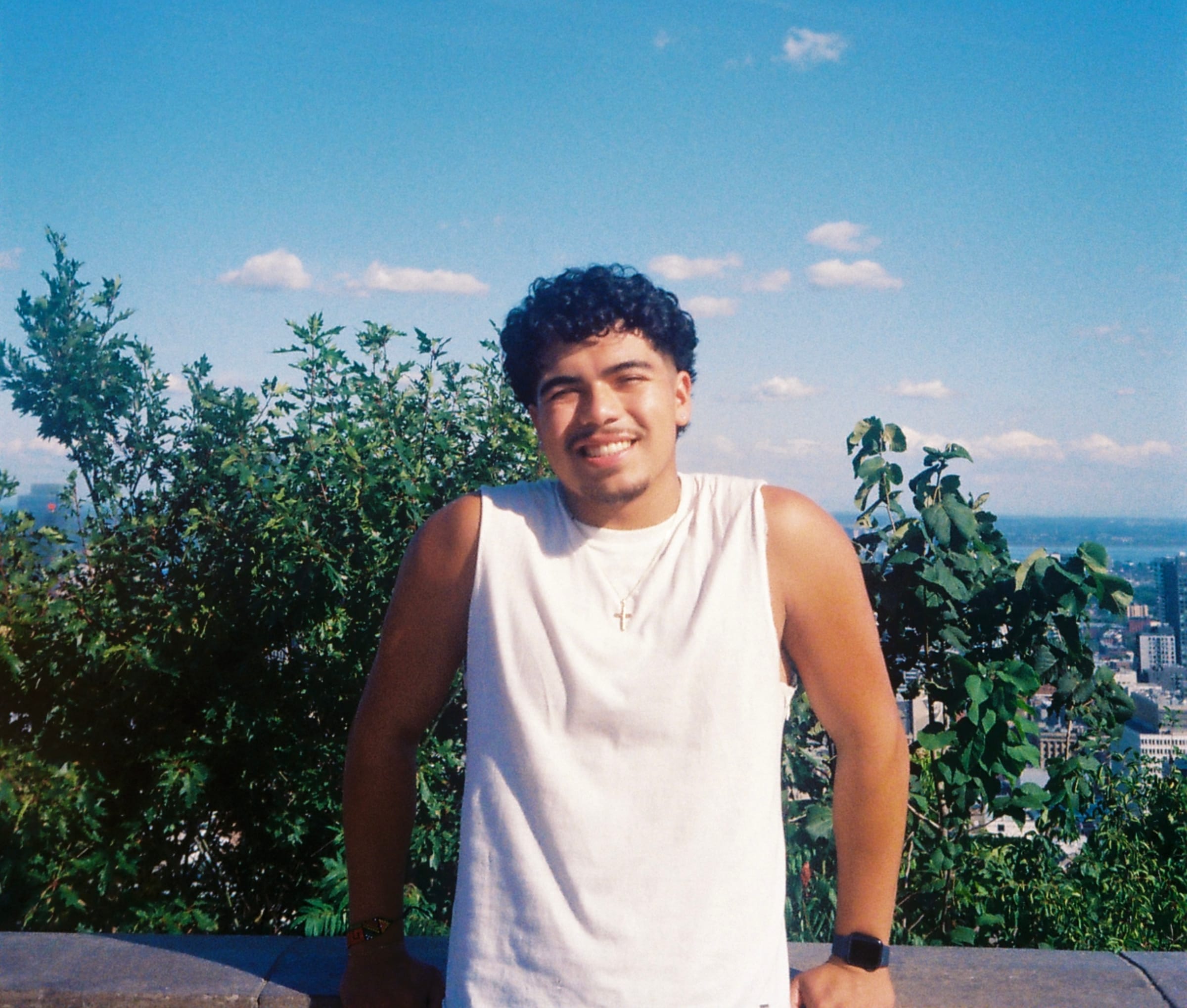 Mateo Burga Villarreal souriant, en débardeur blanc, devant un ciel bleu et la ville en arrière-plan